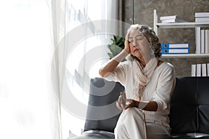 Depressed Asian senior woman sit on sofa looking away by the window. Loneliness elderly female living alone at home with