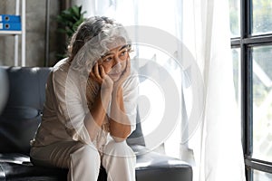 Depressed Asian senior woman sit on sofa looking away by the window. Loneliness elderly female living alone at home with