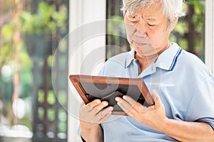 Depressed asian senior woman waiting for the family to visit her with holding a picture frame and watching old photographs of