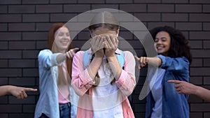 Depressed bullying victim covering face with hands, classmates pointing fingers