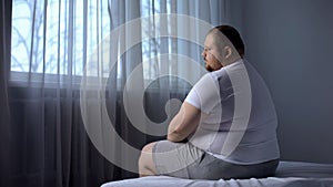 Depressed fat man sitting on bed at home, worried about overweight, insecurities