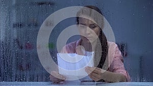 Depressed female reading letter behind rainy window, bad news from relatives