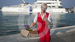 Depressed frustrated Caucasian businesswoman with cardboard box sitting on embankment sighing and thinking. Portrait of