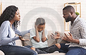 Depressed little black girl sitting between arguing parents, closing ears