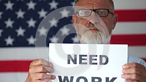 Depressed man with a beard is holding a sign saying need work. Economic crisis metaphor. American flag on the background