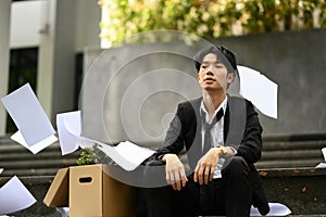 Depressed man office worker sitting on the stairs of building with falling paperwork. Dismissal from work, unemployment