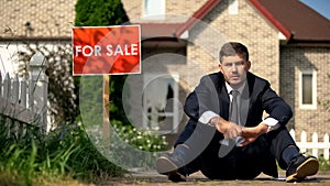 Depressed man sitting on ground near for sale sign, home loss because of debts