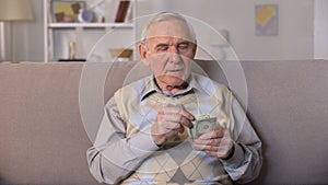 Depressed old man sitting on sofa, holding dollars banknotes, social insecurity