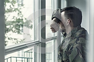 Depressed and sad soldier in green uniform with trauma after war standing near the window photo
