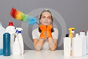 Depressed sad woman looks stressfully, keeps both hands under chin, wearing protective gloves, holding pp duster, surrounded with