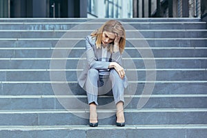 Depressed sad woman sitting on the stairs of a business building, businesswoman in business clothes tired