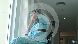 Depressed senior Asian woman patient in hospital sitting in wheelchair by the window, upset disabled patient thinking and looking