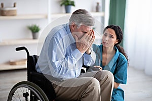 Depressed senior man in wheelchair crying, young doctor trying to help him at retirement home