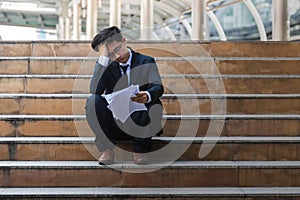 Depressed stressed young Asian business man in suit with hands on head sitting on stairs. Unemployment and layoff concept