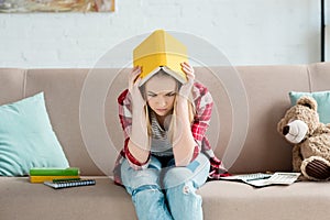 depressed teen student girl with book on head sitting on couch