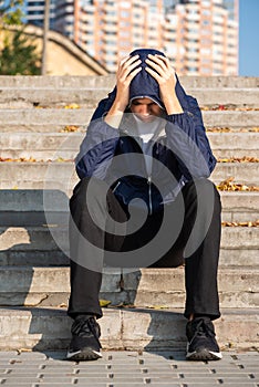 Depressed teenage boy holding his head with his hands sitting on stairs outdoors. Teen depression concept