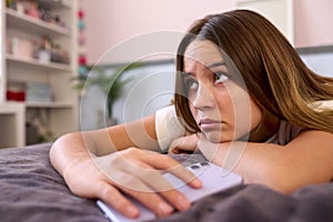 Depressed Teenage Girl Lying On Bed At Home Looking At Mobile Phone