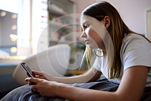 Depressed Teenage Girl Lying On Bed At Home Looking At Mobile Phone