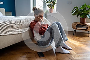 Depressed teenage girl sitting on floor at home with smartphone waiting for call from boyfriend