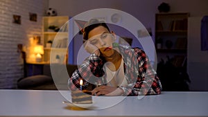 Depressed teenager celebrating birthday, looking at cake with candle, outsider