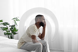 Depressed unhappy tired young black man sits on bed and think in bedroom interior, side view