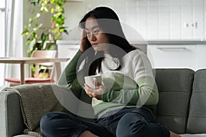 Depressed weakened Asian woman millennial with cup of coffee sits on sofa in living room of home
