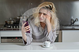 Depressed woman in the kitchen in the morning. Upset woman reading message or bad new at smartphone in the kitchen. Sad