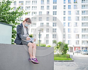 Depressed woman in medical mask sits on parapet outdoors with a box of personal items from the desktop. Unemployed girl