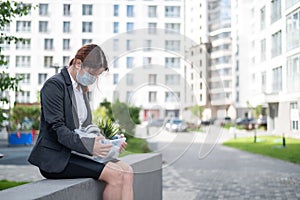 Depressed woman in medical mask sits on parapet outdoors with a box of personal items from the desktop. Unemployed girl