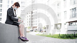 Depressed woman in medical mask sits on parapet outdoors with a box of personal items from the desktop. Unemployed girl