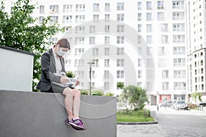Depressed woman in medical mask sits on parapet outdoors with a box of personal items from the desktop. Unemployed girl