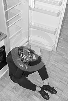 Depressed Woman Sitting on the Floor in Front of an Empty Refrigerator at Home