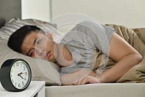 Depressed young Asian man lying in bed cannot sleep from insomnia, focus on alarm clock