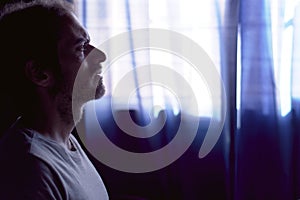 Depressed young bearded man crying in dark room. Dim light with window behind. Concept of depression and mental health, grief and