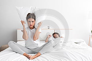 Depressed young caucasian lady sits on bed covers her ears with pillow and suffers from snoring of her sleeping husband