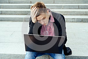 Depressed young handsome man sitting outdoors and looking at laptop.