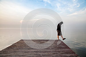 Depressed young man wearing a black hoodie standing on wooden bridge extended into the sea looking down and contemplating suicide