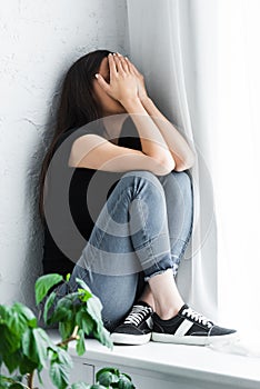 depressed young woman crying while sitting on window sill and covering face with hands.