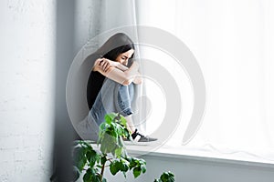 depressed young woman crying while sitting on window sill and hiding face in crossed arms.
