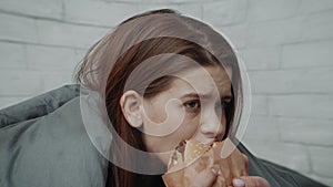 Depressed Young Woman Eats Burger Comforting Herself Sitting In Bedroom