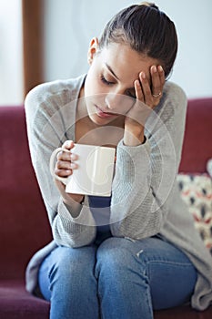 Depressed young woman having headache while drinking coffee on sofa at home