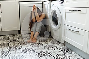 Depressed young woman or teenager sitting on floor in kitchen corner with hands close face.