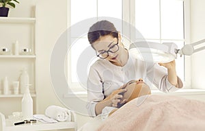 Dermatologist, skin therapist, or beautician examining skin on woman& x27;s face under magnifying lamp