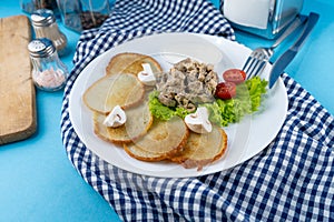 Deruny - potato pancakes with mushrooms, champignons, sour cream on a white plate. Cherry tomatoes, salad. Restaurant serving on a