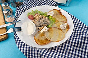 Deruny - potato pancakes with mushrooms, champignons, sour cream on a white plate. Cherry tomatoes, salad. Restaurant serving on a