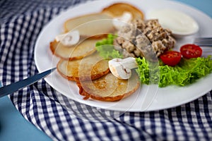 Deruny - potato pancakes with mushrooms, champignons, sour cream on a white plate. Cherry tomatoes, salad. Restaurant serving on a