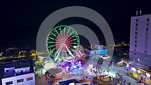 descending aerial footage of of the Carolina Beach Boardwalk with a Ferris wheel and colorful amusement rides and games, people