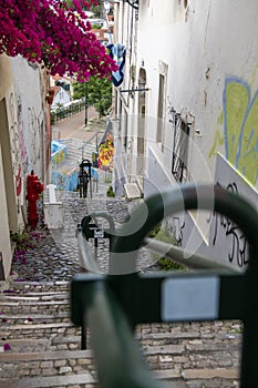 Descent of stone stairs between houses with red hanging flowers with central green metal railing