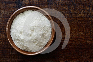 Desiccated coconut in dark wooden bowl isolated on dark brown wood from above.