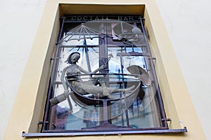 Design of a window grill with a stone girl drinking a cocktail while relaxing inside a cup-shaped sofa below a parasol, in Cesky